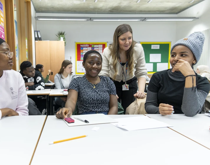 A classroom scene where three female students are smiling and engaging in discussion. A teacher in a beige cardigan leans over the table to interact with them, while other students work in the background. A classroom scene where three female students are smiling and engaging in discussion. A teacher in a beige cardigan leans over the table to interact with them, while other students work in the background.